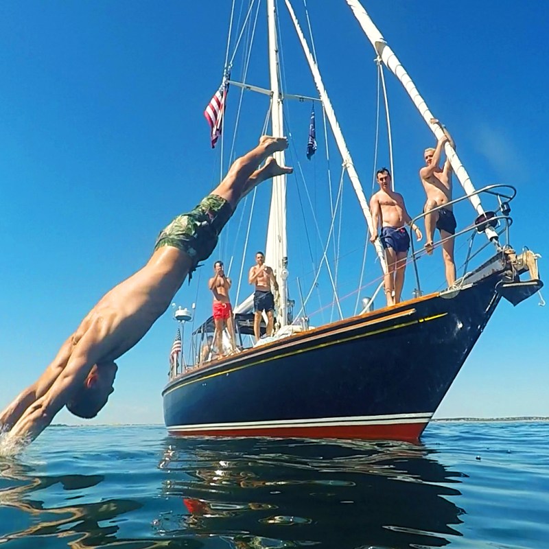 Group of men standing on sailboat with close up of man diving into water from bow of boat