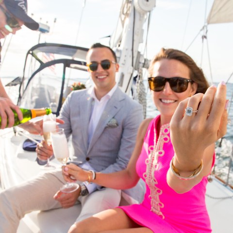 Women holding up hand to show off engagement ring with fiance behind her on sailboat