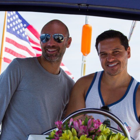 Two men smiling at camera with American flag waving behind them