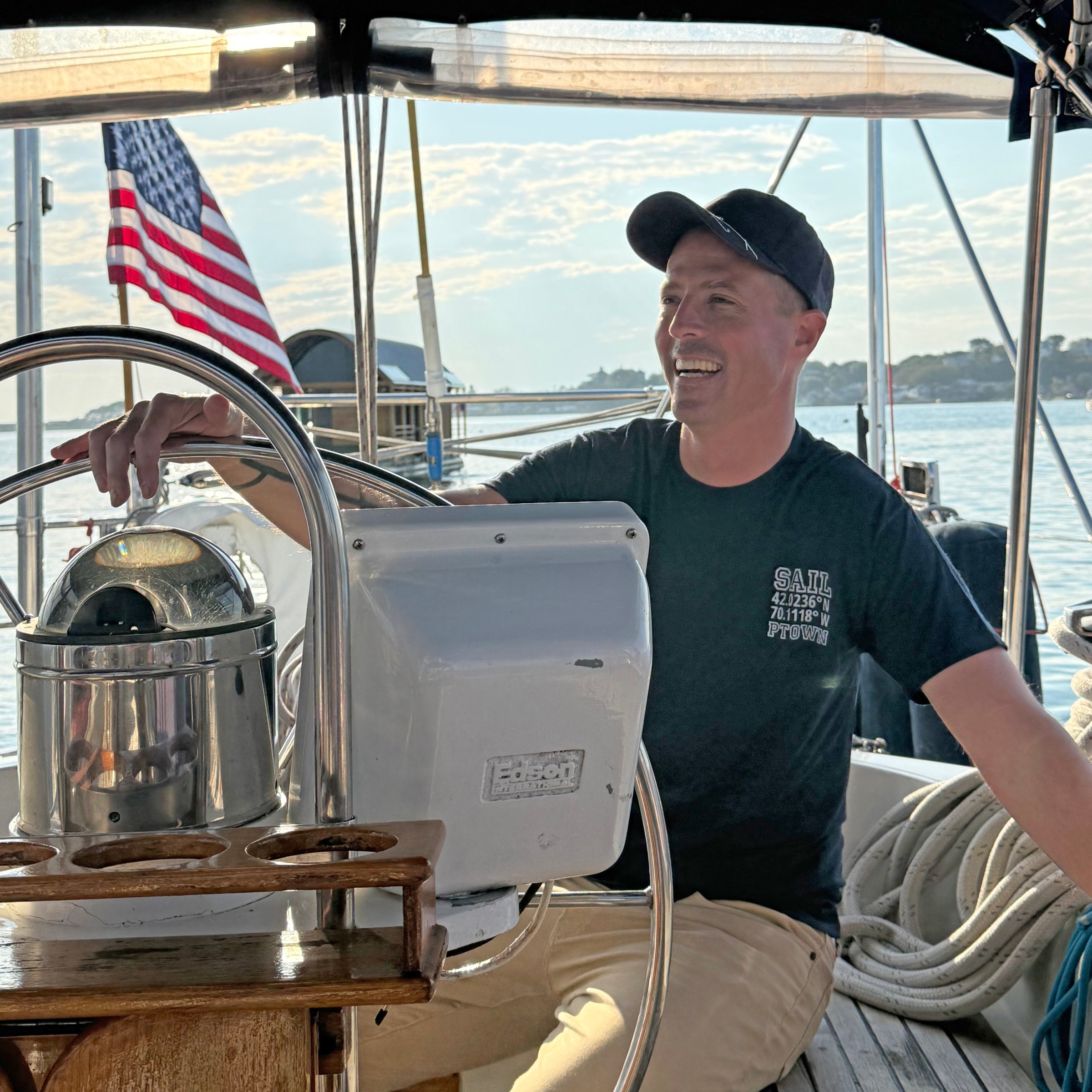 Smiling person steering a boat with an American flag in the background.