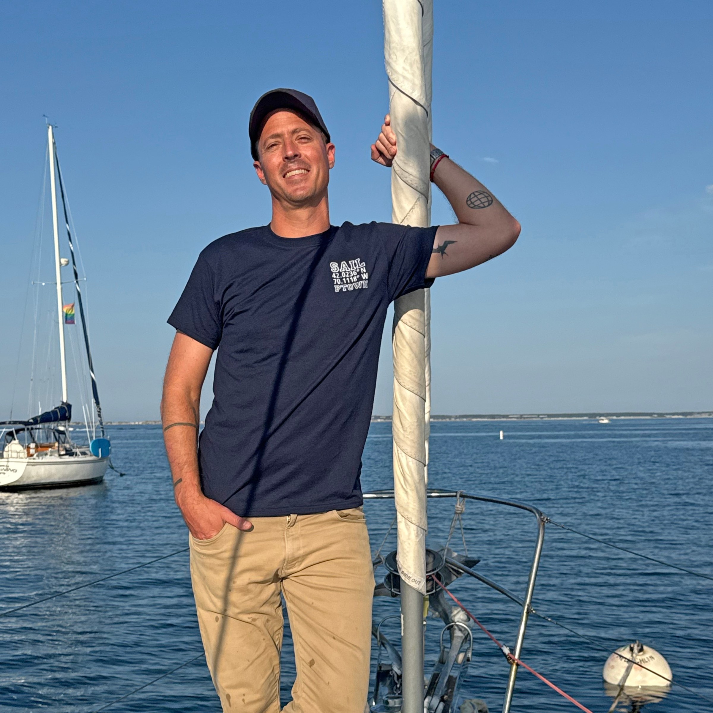 Person in navy shirt and cap leaning on sailboat mast, smiling, with another sailboat in the background.